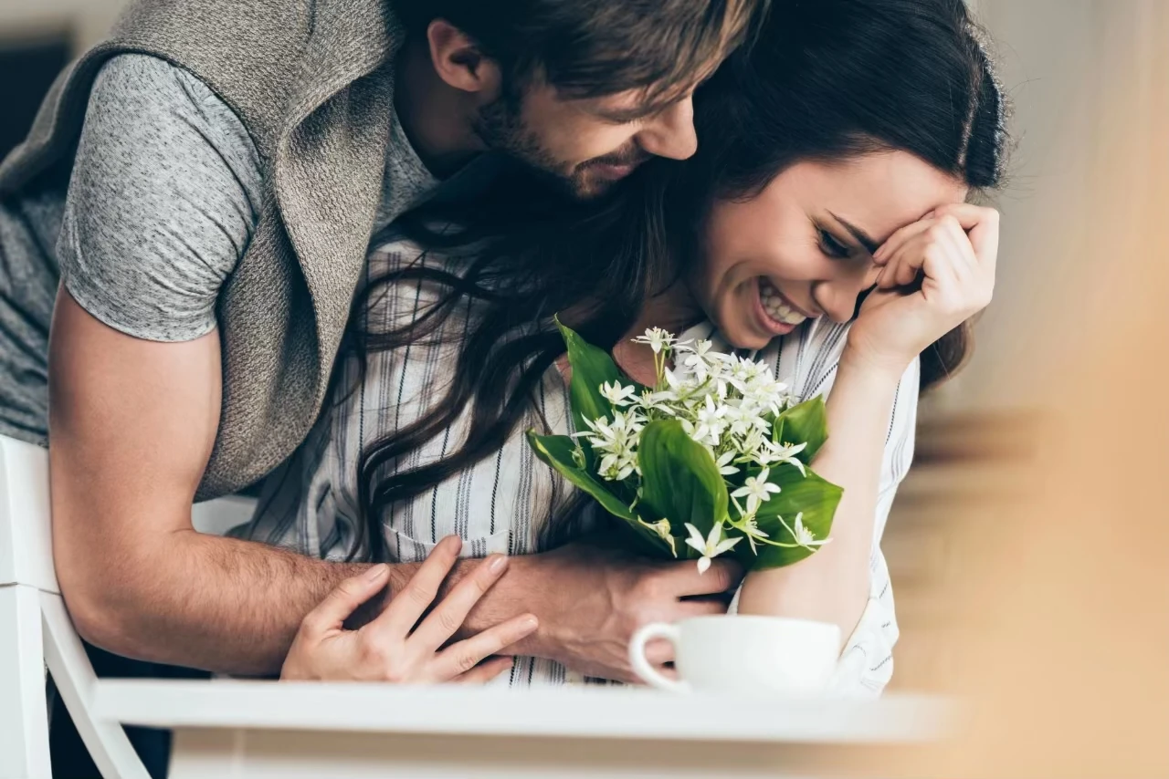 Frau sitzt am Tisch, Mann beugt sich über sie mit Blumenstrauß mit weißen Blumen in der Hand; sie lächelt