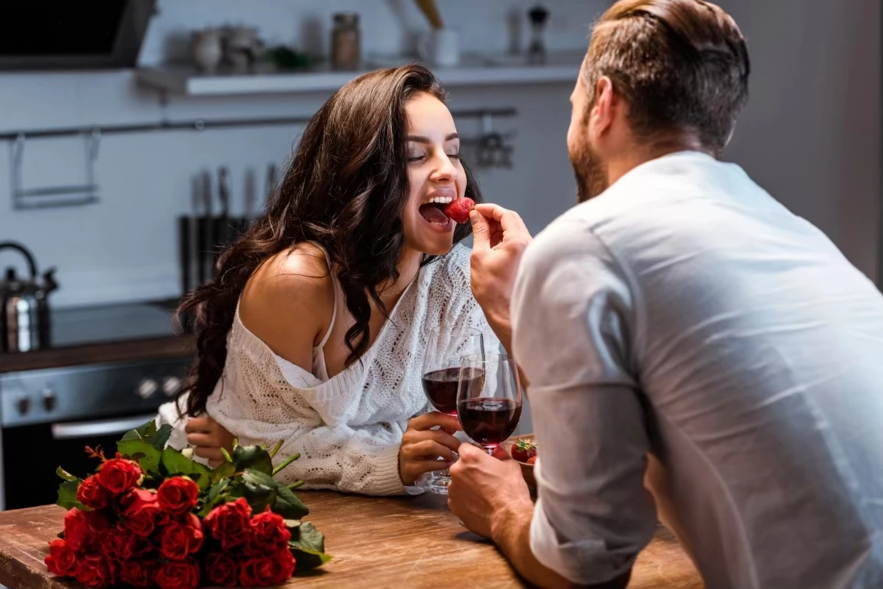 Frau und Mann lehnen über Küchentresen, neben ihnen ein Rosenstrauß, er füttert ihr eine Erdbeere