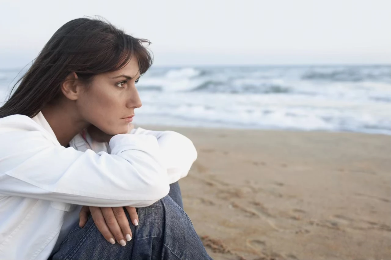 Frau sitzt am Meer, die Beine angezogen, die Arme auf den Knien, mit traurigem Blick aufs Meer