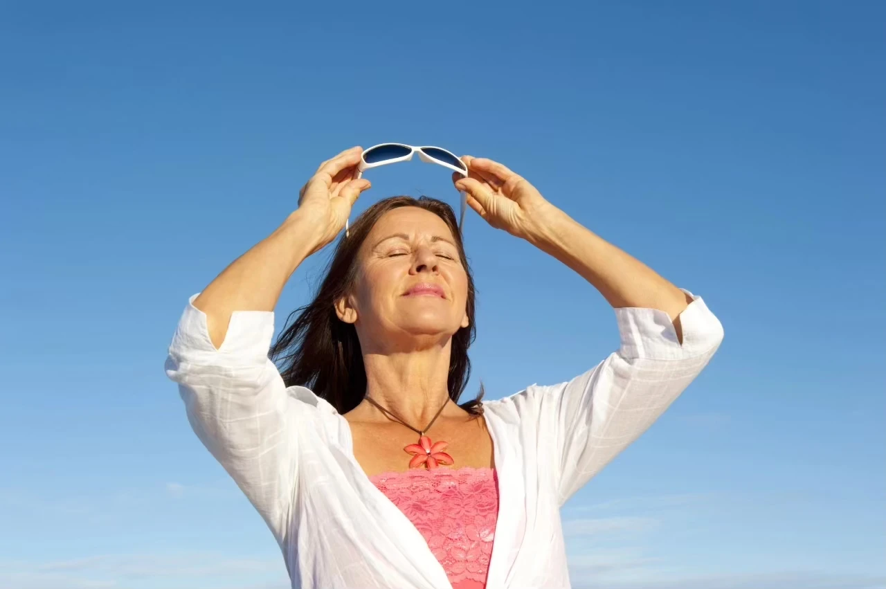 Frau in pinkfarbenem Top und weißer Bluse vor blauem Himmel hat die Augen entspannt geschlossen und setzt sich eine Sonnenbrille auf