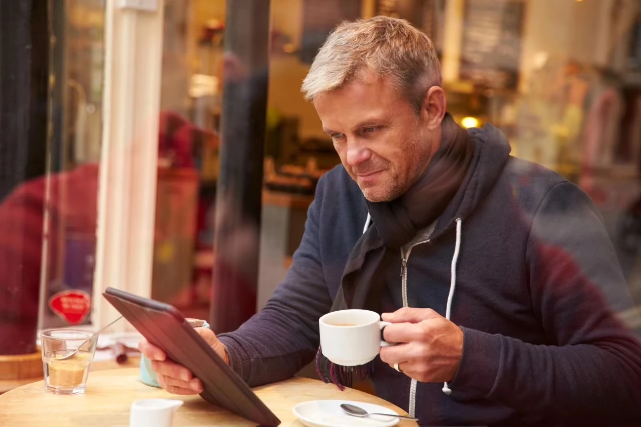 Mann sitzt mit gefrorenem Lächeln in Café, Tasse in der einen Hand, Tablet in der anderen