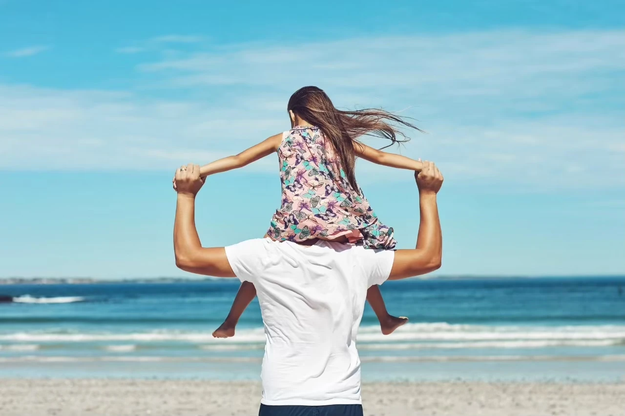 Erwachsene Person am Strand mit Blick aufs Meer trägt Kind auf den Schultern