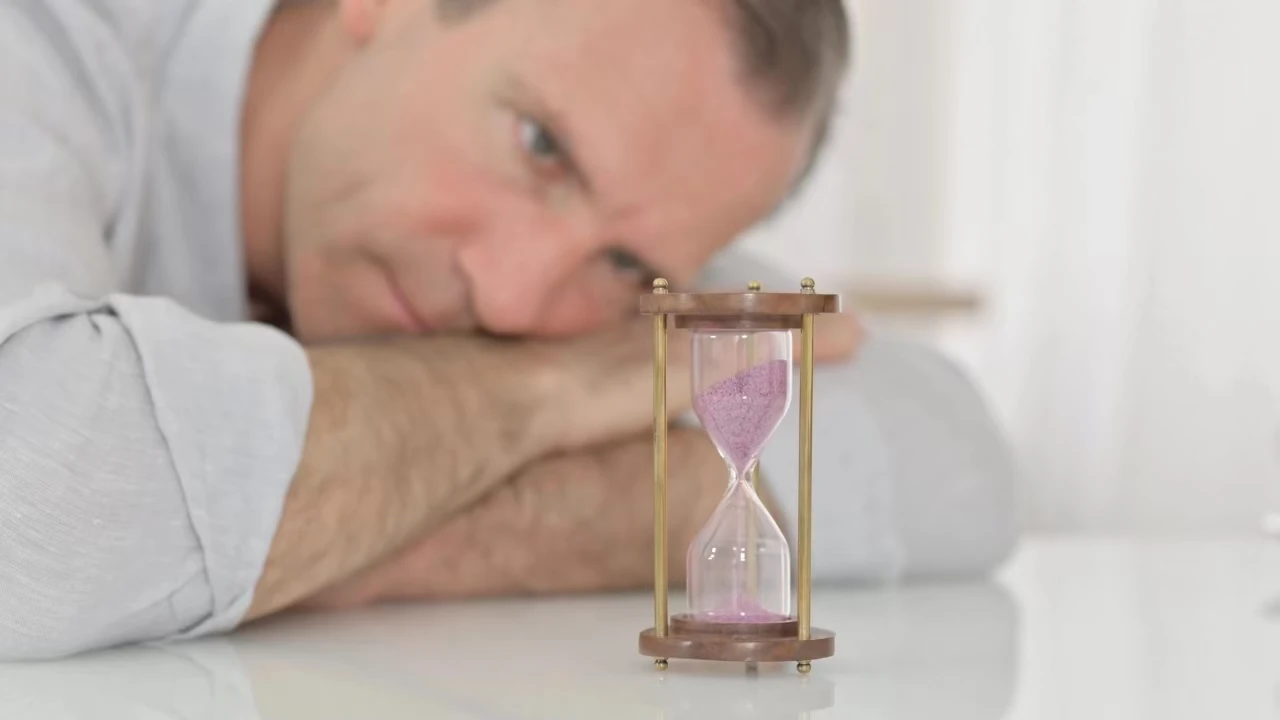 Mann liegt mit dem Kopf auf seinen Armen am Tisch und beobachtet, wie Sand durch eine Sanduhr rieselt