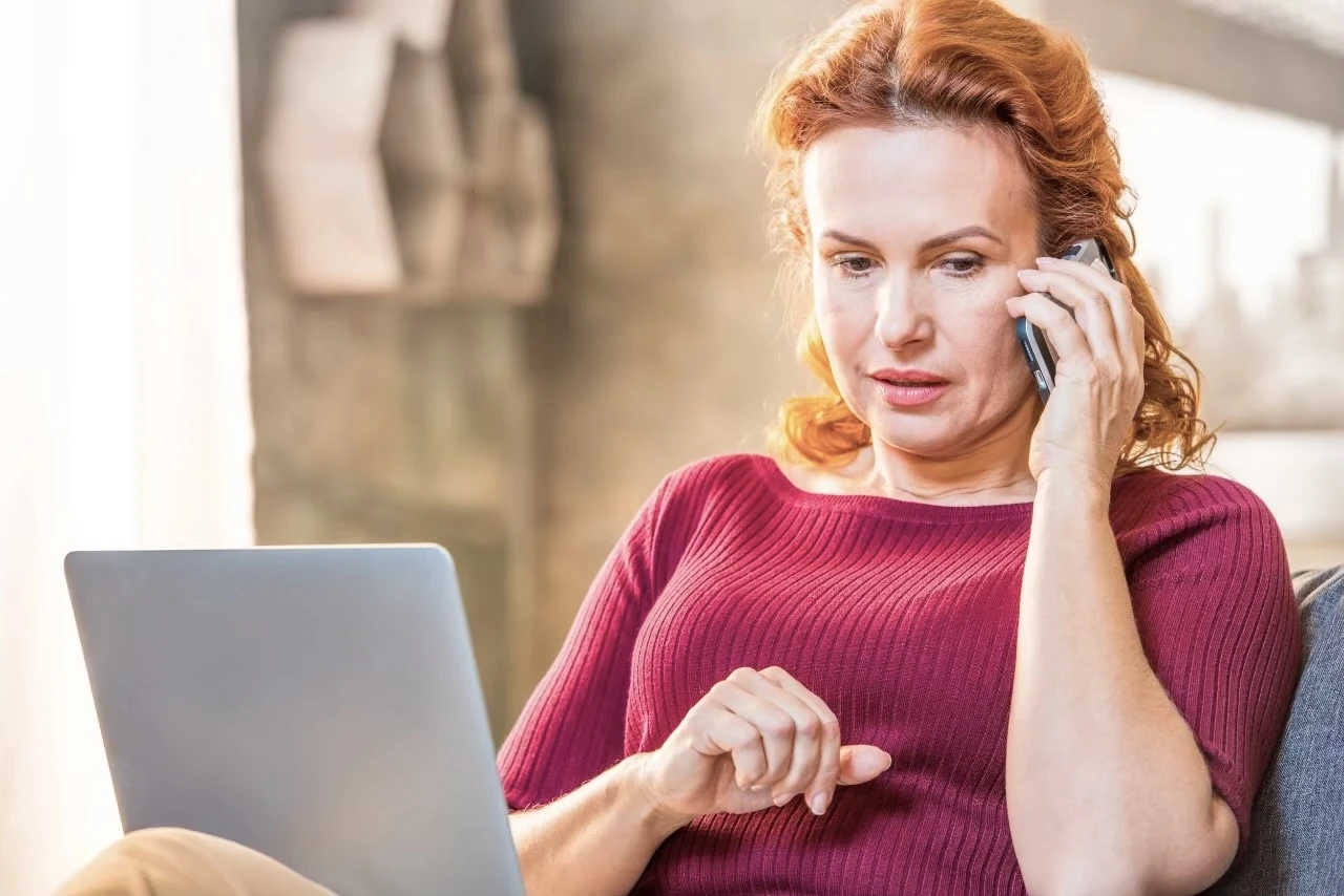 Frau sitzt mit Laptop auf den Knien auf dem Sofa und telefoniert mit dem Handy
