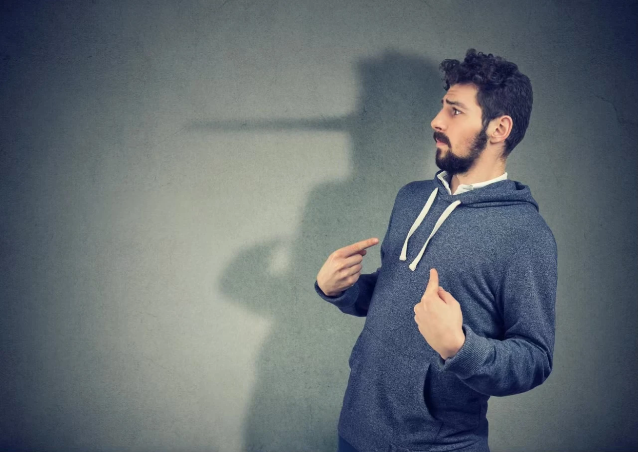 Mann lehnt sich zurück, zeigt mit beiden Händen ungläubig auf sich; an der Wand ein Schatten, die Nase langgezogen wie die von Pinocchio