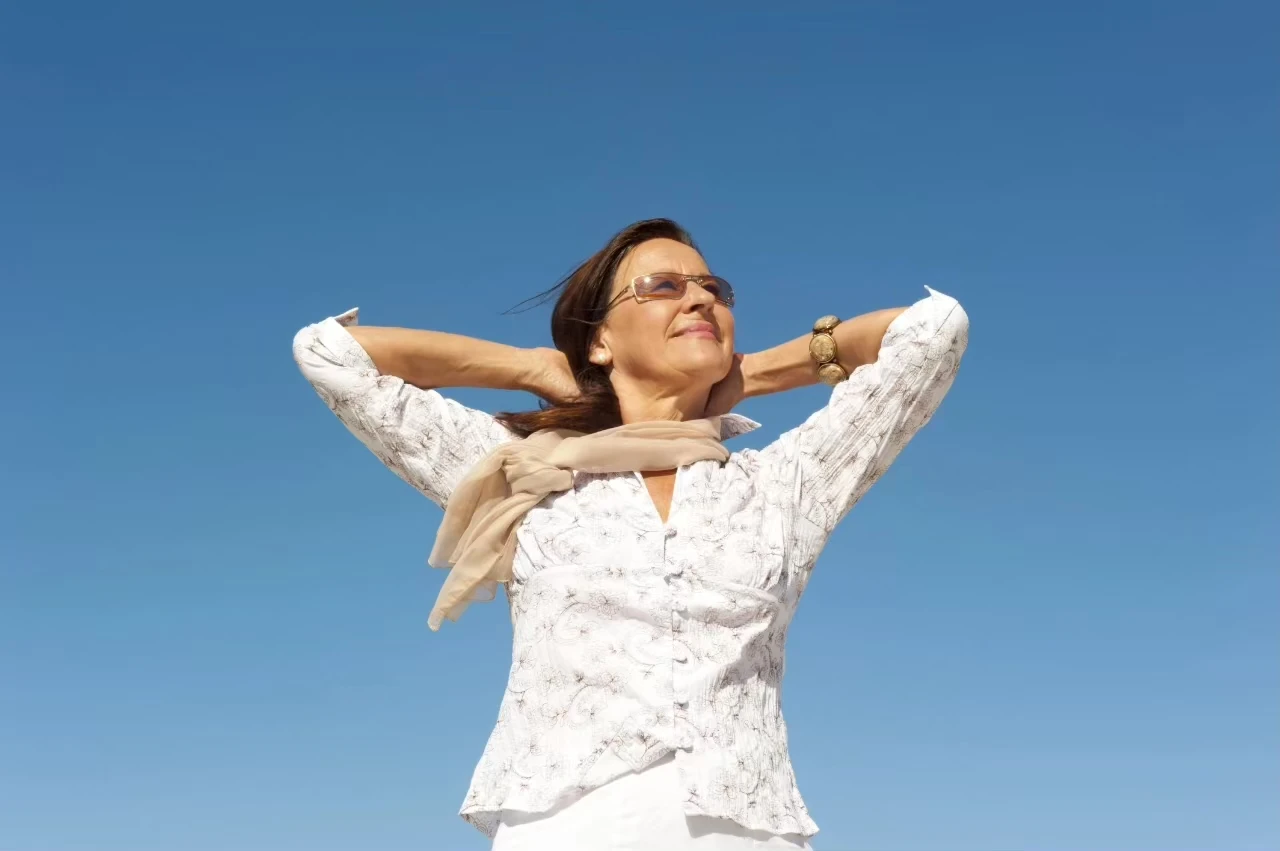 Frau mit weißer Bluse und Sonnenbrille hat die Hände verschränkt und lächelt, hinter ihr blauer Himmel