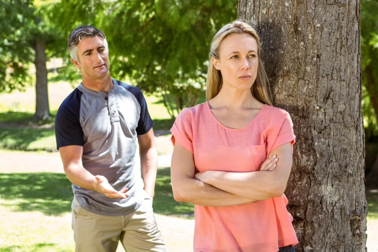 Frau steht mit verschränkten Armen an einem Baum, hinter ihr ein Mann, der die Hand nach ihr ausstreckt