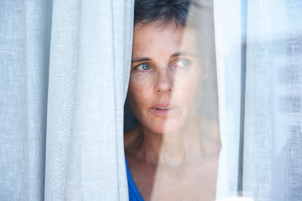 Eine Person schaut zögerlich/ängstlich durch Vorhänge aus dem Fenster