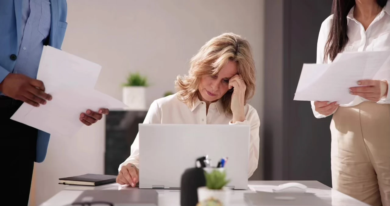 Frau schaut auf Laptop vor ihr, den Kopf auf eine Hand gestützt, rechts und links von ihr zwei Personen mit Dokumenten